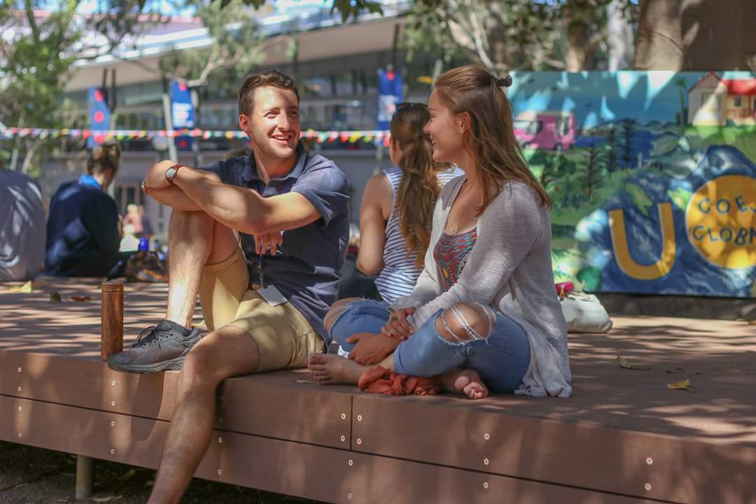 Zwei Studierende sitzen auf einer Mauer am Campus